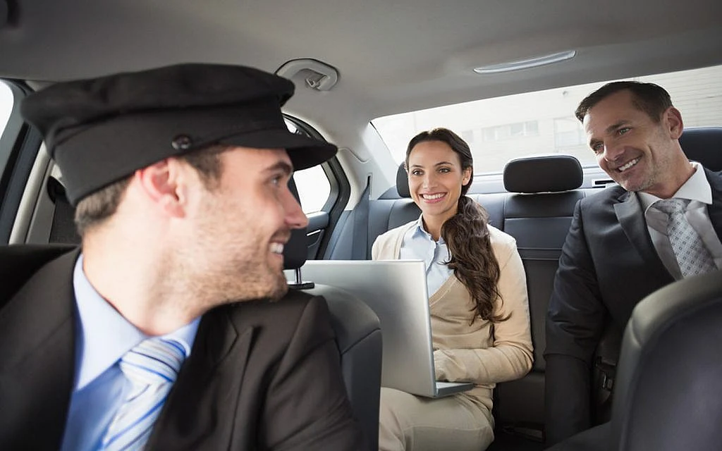 Business professionals smiling inside a car, with a chauffeur driving, man and woman in back seat, using a laptop.